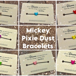May include: A set of six Mickey Mouse themed bracelets with different colored charms. Each bracelet has a black string and a charm shaped like Mickey Mouse's head. The charms are in different colors, including gold, purple, blue, yellow, green, and pink. The bracelets are displayed on cards with the quote "All our DREAMS can come true if we have the courage to pursue them. - Walt Disney".