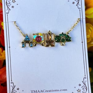 May include: Gold-toned necklace featuring charm pendants shaped like a castle, a large iridescent gem, a building, and a tree with green gems. The necklace is displayed on a white card with the website TMMAACreations.com.