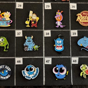 May include: A collection of colorful enamel pins featuring various cartoon characters. Designs include characters from Disney movies, such as Mike Wazowski, Sully, Dory, and Genie. The pins are displayed on a black grid.