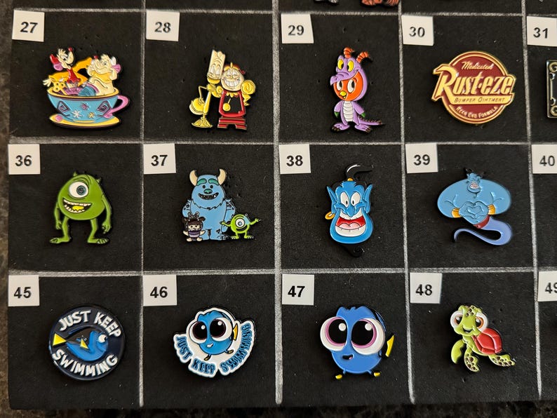 May include: A collection of colorful enamel pins featuring various cartoon characters. Designs include characters from Disney movies, such as Mike Wazowski, Sully, Dory, and Genie. The pins are displayed on a black grid.