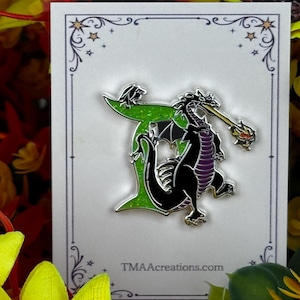 May include: A metal enamel pin featuring a black dragon with a purple underbelly, breathing fire, positioned in front of a glittery green letter T. The pin is attached to a white display card with the text TMAcreations.com printed at the bottom.