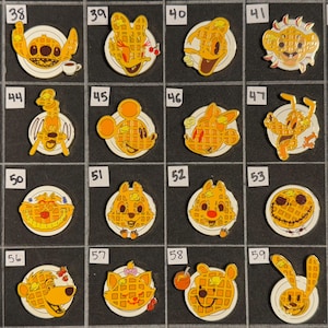 May include: A collection of enamel pins featuring various cartoon characters designed as waffles on plates. The pins include characters like Stitch, Mickey Mouse, Donald Duck, and Winnie the Pooh. Each pin is a different character.