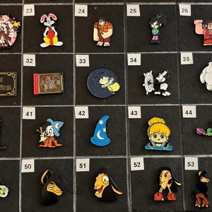 May include: A collection of colorful enamel pins featuring various cartoon characters and themed designs. Includes pins of a rabbit, a wizard hat, a book, and characters from animated films. The pins are arranged on a black surface.