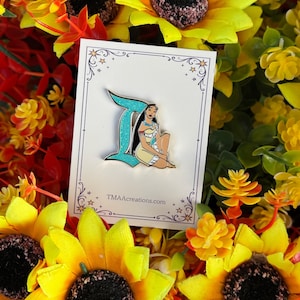 May include: A metal enamel pin featuring a glittery teal letter I and a cartoon character with long black hair wearing a tan dress. The pin is attached to a white rectangular display card with decorative star borders and the text TMAcreations.com.
