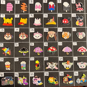 May include: A collection of colorful enamel pins featuring various cartoon characters and objects. Designs include ice pops, character heads, umbrellas, popcorn, and vehicles. The pins are arranged on a black surface.