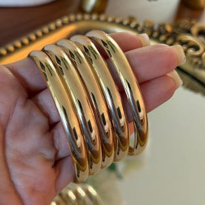 May include: A hand holds a stack of six gold-colored, polished bangles. The bangles are thick and rounded, reflecting light. The background includes a gold-framed mirror and other decorative items.