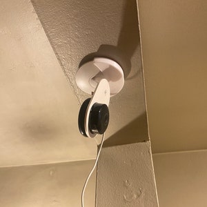 Op de afbeelding: Een witte beveiligingscamera gemonteerd op een witte plafondbevestiging. De camera is verbonden met een wit snoer.