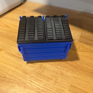 May include: A miniature blue and black dumpster model. The body is a vibrant blue, while the lid and top section are black. The model is designed with realistic details, including a hinged lid and textured surfaces.