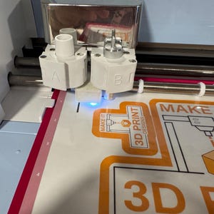 May include: A close-up of a white and blue 3D printer cutting a design. The machine has two white heads labeled "A" and "B". The design is orange and black and says "3D PRINT DESIGNS" and "MAKE IT".