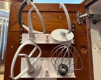 Op de afbeelding: Een set van vier witte keukenmixer-accessoires die aan een houten kastdeur hangen. De accessoires omvatten een deeghaak, een platte klopper, een garde en een spiraalhaak.