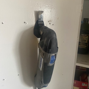 May include: A gray and blue Dremel multi-tool with a black blade attached. The tool is mounted on a white wall with a black plastic holder.