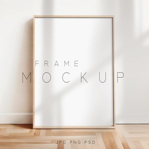 以下が含まれることがあります： 白い背景に黒い文字で「FRAME MOCKUP」と書かれたシンプルな木製フレームのモックアップ。フレームは明るい木製の床に置かれ、背景には白い壁がある。