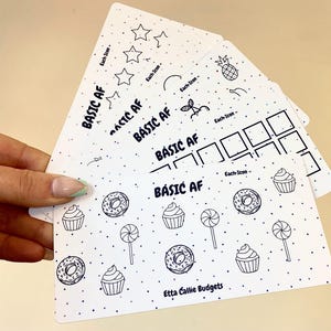 May include: A set of five white cards with black ink, each featuring a different design. The cards are labelled "BASIC AF" and feature icons such as stars, a pineapple, a plant, a cupcake, a donut, and a lollipop.