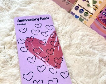 Puede incluir: Una tarjeta lila con el texto "Anniversary Funds" y "Each Icon -" presenta corazones negros delineados y un gran corazón rosa. La tarjeta está sobre una superficie blanca y esponjosa. Otras tarjetas con corazones y números son visibles en el fondo.