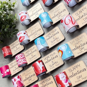 Peut inclure: Un ensemble de bracelets à claquer sur le thème de la Saint-Valentin avec différents motifs et messages colorés. Les bracelets présentent des phrases comme "Oh Snap! Happy Valentine's Day" et "Hope Your Valentine's is Slappin'" avec différents noms d'expéditeurs.