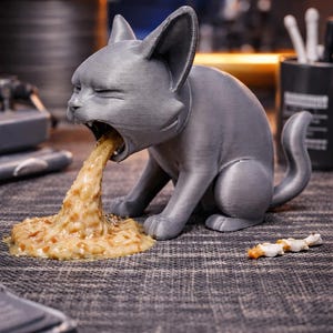 May include: A gray cat figurine in a seated position, mid-vomit, with a stream of beige and brown material emanating from its mouth. A small, white and brown object lies nearby. The cat has closed eyes and a curled tail.
