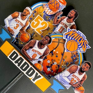 May include: A basketball-themed cake topper featuring New York Knicks players in action. The design includes the team logo, basketballs, a basketball hoop, and the word "DADDY". The color scheme is orange, blue, and white.