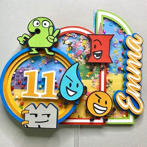 May include: A colorful cake topper featuring cartoon characters and the number 11. The design includes a blue and yellow circular frame, a red frame, and the name "Emma" in orange script. The topper is decorated with confetti.