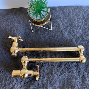 Puede incluir: Un porta ollas de doble brazo de metal dorado con un diseño giratorio. El porta ollas está montado en la pared y tiene dos brazos que se extienden hacia afuera. Los brazos están conectados por una barra de metal. El porta ollas está hecho de metal pulido y tiene un acabado brillante.