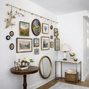 May include: A gallery wall featuring a variety of framed art pieces, including paintings and prints, suspended from a gold-toned rod. A round wooden table with candles and a clock is in the foreground, with a marble-topped table and mirror nearby.