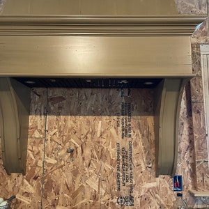 Puede incluir: Una campana de cocina de madera marrón con un borde decorativo y dos luces. La campana está montada en una pared con madera contrachapada sin terminar.