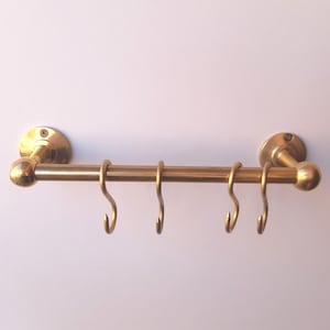 Puede incluir: Un riel de cocina de color latón con cuatro ganchos en forma de S. El riel está montado en una pared blanca y tiene extremos redondeados decorativos. Los ganchos están espaciados uniformemente a lo largo de la barra horizontal.