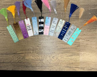 Puede incluir: Una colección de marcapáginas coloridos con borlas. Cada marcapáginas presenta un diseño y texto únicos, incluyendo frases como "A lot to read at the moment" y "Life of a sun". Las borlas vienen en varios colores, como amarillo, morado y azul.