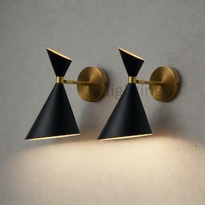 Könnte beinhalten: Zwei schwarze, kegelförmige Wandleuchten mit messingfarbenen Montageplatten und Armen. Die Leuchten haben ein modernes Design mit einer dreieckigen Oberseite und einer nach unten gerichteten Lichtquelle. Sie sind an einer hellgrauen Wand montiert.