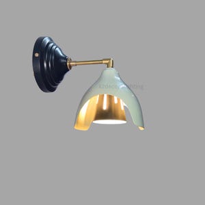 Puede incluir: Una lámpara de pared con una base negra y una pantalla blanca curva con un interior dorado. La pantalla tiene un diseño único con una sección recortada, que revela la bombilla.