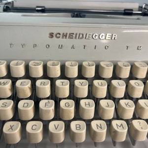 May include: A vintage gray typewriter with the brand name "SCHEIDEGGER TYPOMATIC TMS" printed on the top. The keyboard has keys for letters, numbers, and symbols.