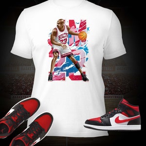 Könnte beinhalten: Weißes T-Shirt mit einem Aufdruck von Michael Jordan, der ein Trikot der Chicago Bulls trägt und einen Basketball dribbelt. Der Aufdruck ist in einem rot-blau-weißen Farbschema gehalten. Das T-Shirt wird mit einem Paar schwarz-roter Sneakers kombiniert.