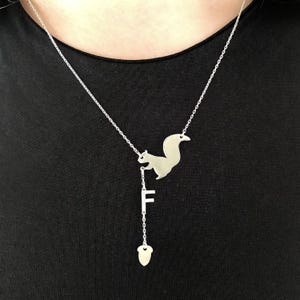 Könnte beinhalten: Eine silberfarbene Halskette mit einem Eichhörnchen-Anhänger, dem Buchstaben "F" und einem Eichel-Charm. Die Halskette hängt an einer feinen Kette und erzeugt ein skurriles Design. Der Anhänger ist detailliert und hat eine polierte Oberfläche.