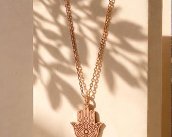 May include: A rose gold-toned necklace featuring a hamsa hand pendant. The pendant displays an engraved eye motif at the centre, surrounded by intricate floral and geometric line patterns. The piece hangs from a delicate, thin metal link chain.