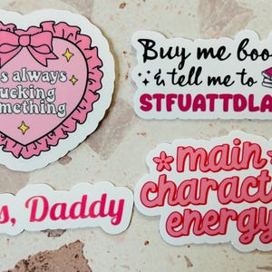 May include: Four stickers with text. One pink heart-shaped sticker with the text "It's always fucking something". Another sticker says "Buy me books & tell me to STFUATTDLAGG". A third sticker says "Yes, Daddy", and the last says "main character energy".