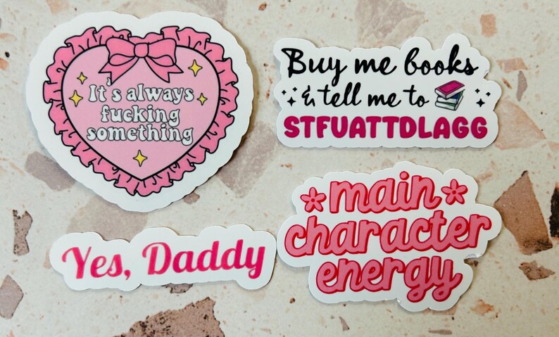 May include: Four stickers with text. One pink heart-shaped sticker with the text "It's always fucking something". Another sticker says "Buy me books & tell me to STFUATTDLAGG". A third sticker says "Yes, Daddy", and the last says "main character energy".