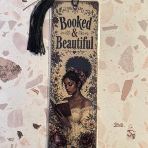 Op de afbeelding: Een rechthoekige boekenlegger met een zwarte kwast. De boekenlegger toont een illustratie van een vrouw die een boek leest, omringd door bloemmotieven. De tekst "Booked & Beautiful" staat bovenaan.