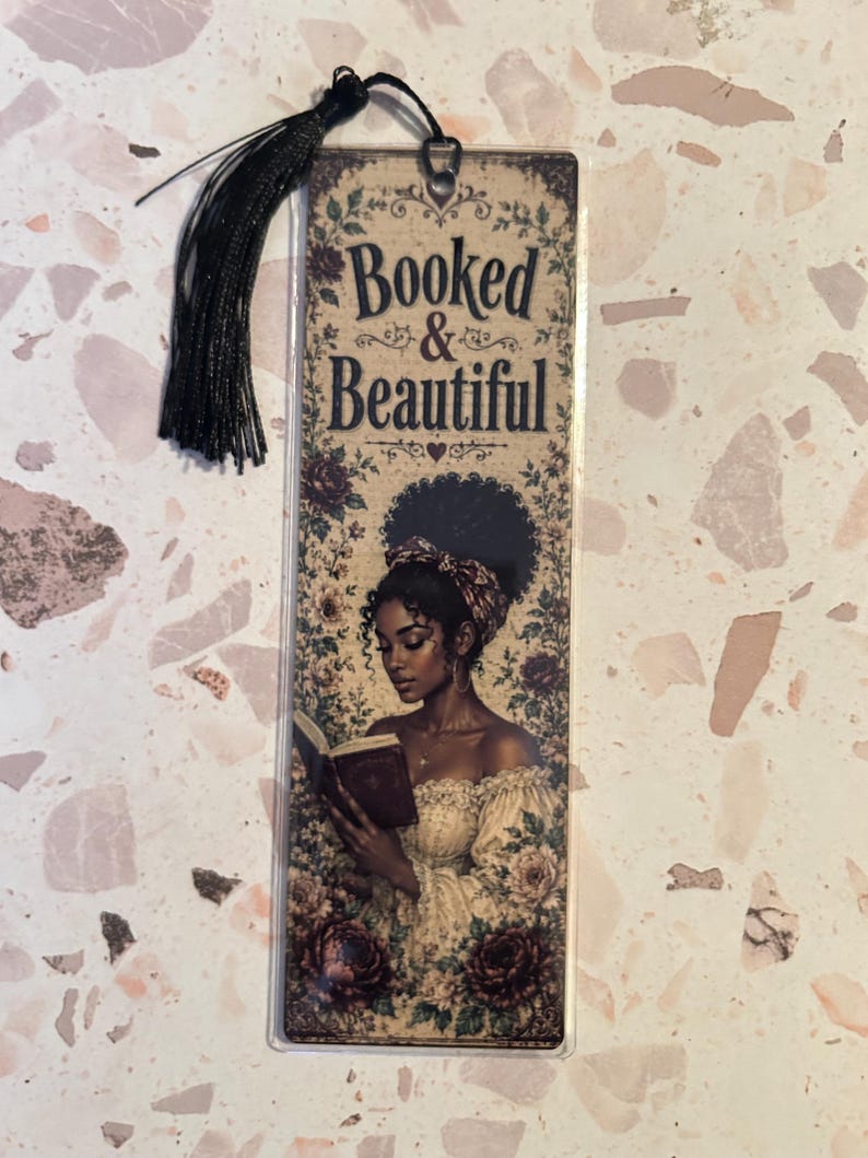 Op de afbeelding: Een rechthoekige boekenlegger met een zwarte kwast. De boekenlegger toont een illustratie van een vrouw die een boek leest, omringd door bloemmotieven. De tekst "Booked & Beautiful" staat bovenaan.