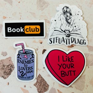 May include: A collection of stickers with various designs. One sticker reads "Book club" in white text on a black rectangle with an orange accent. Another features a drawing of a person on a book with the text "STFUATTDLAGG". A heart-shaped sticker says "I Like YOUR BUTT".