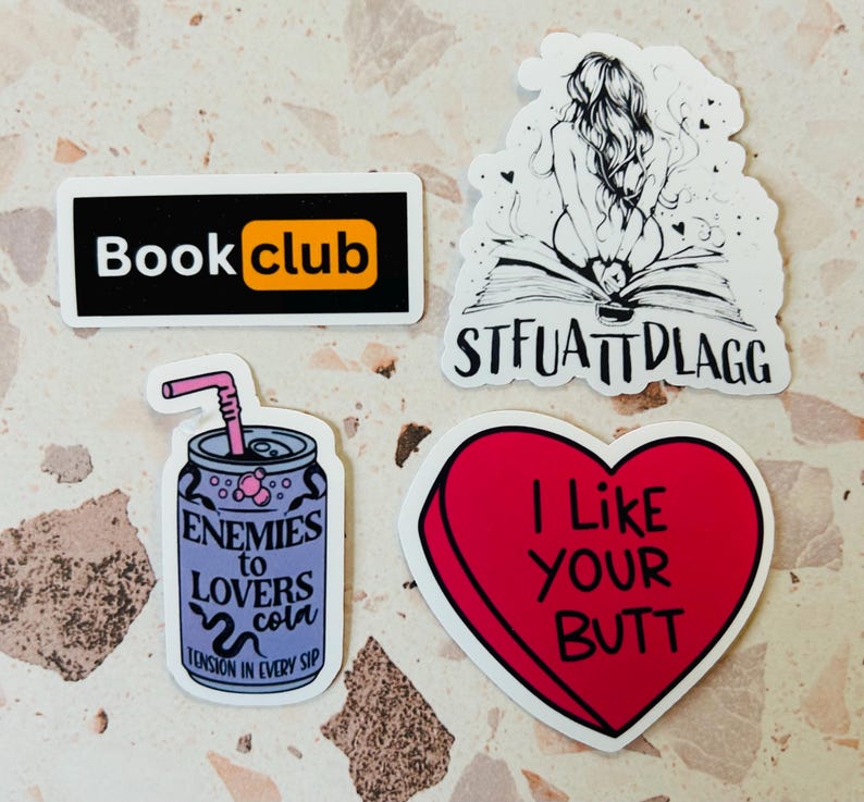 May include: A collection of stickers with various designs. One sticker reads "Book club" in white text on a black rectangle with an orange accent. Another features a drawing of a person on a book with the text "STFUATTDLAGG". A heart-shaped sticker says "I Like YOUR BUTT".