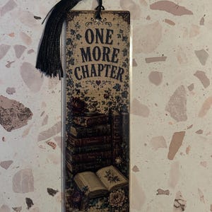 Op de afbeelding: Een rechthoekige boekenlegger met de tekst "ONE MORE CHAPTER" in vintage stijl. De boekenlegger heeft een stapel boeken, bloemmotieven en een zwarte kwast. De achtergrond is beige.