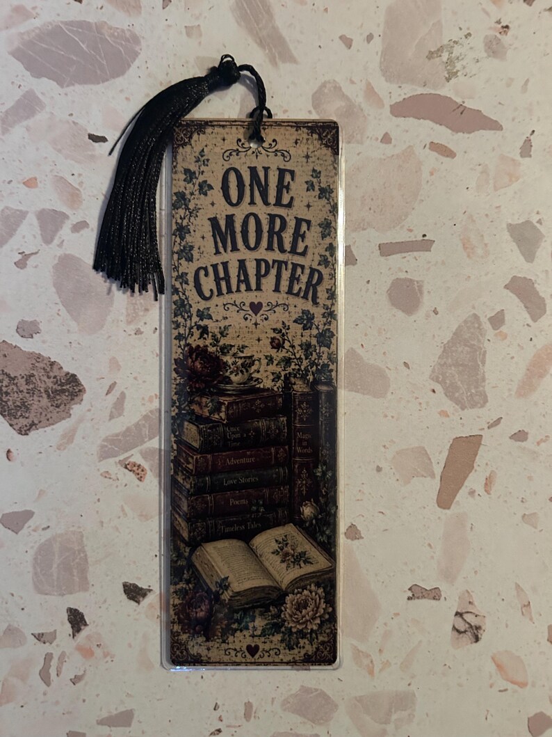 Op de afbeelding: Een rechthoekige boekenlegger met de tekst "ONE MORE CHAPTER" in vintage stijl. De boekenlegger heeft een stapel boeken, bloemmotieven en een zwarte kwast. De achtergrond is beige.