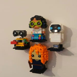 Op de afbeelding: Een set van vier LEGO minifiguren, waaronder Wall-E, Eve, een vrouwelijk personage met oranje haar en een vrouwelijk personage met zwart haar en een groene bril. De minifiguren worden getoond op kleine zwarte standaards.