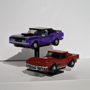 Op de afbeelding: Twee speelgoedauto's, een paarse muscle car met een zwarte motorkap en een rode sportwagen, worden op een standaard getoond. De paarse auto heeft de tekst "442" aan de zijkant. De rode auto heeft de tekst "YNJ197" aan de voorkant.