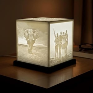 May include: A white, cube-shaped lamp with a black base. The lamp features etched images of an elephant and a group of people. The lamp is lit, casting a warm glow. The lamp is sitting on a wooden surface.