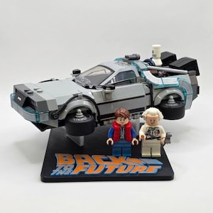 May include: A Lego set featuring a silver DeLorean time machine with black wheels and clear accents, along with two Lego figures. The base is black with the words "BACK TO THE FUTURE" in orange and blue.