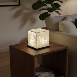 May include: A cube-shaped lamp with images of a rabbit and a puppy, illuminated from within. The lamp sits on a dark base and is placed on a wooden side table. The lamp's design includes paw prints and text.