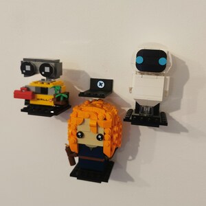 Op de afbeelding: Drie Lego minifiguren die op zwarte planken zijn gemonteerd aan een witte muur. De minifiguren zijn Wall-E, Eve van Wall-E en een roodharig meisje met een blauwe jurk.