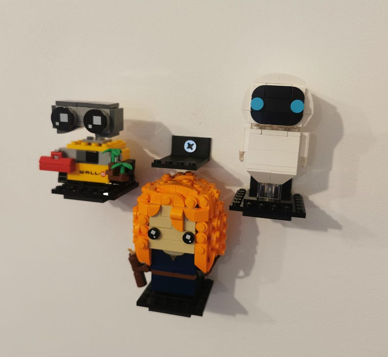 Op de afbeelding: Drie Lego minifiguren die op zwarte planken zijn gemonteerd aan een witte muur. De minifiguren zijn Wall-E, Eve van Wall-E en een roodharig meisje met een blauwe jurk.