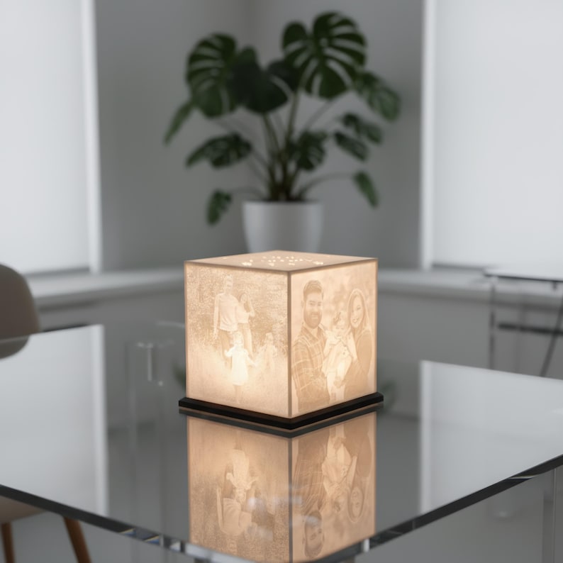 May include: A square lamp with family photos on each side, resting on a black base. The lamp is illuminated, showcasing the images. The lamp is sitting on a glass table, with a plant in the background.