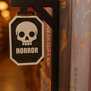 Op de afbeelding: Zwart en wit "HORROR" bord met een schedelafbeelding, gemonteerd op een zwarte metalen beugel. Het bord is geplaatst tussen boeken. Dit is een decoratief item voor een boekenplank.